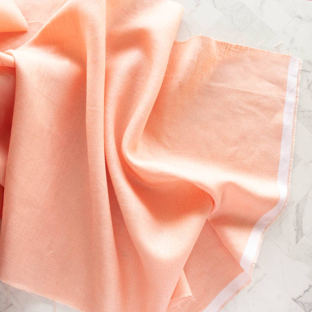 A piece of peach-coloured yarn-dyed linen fabric with a visible white selvedge, displayed on a marble surface.